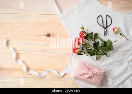 Rosa Rosen, Geschenk in Tuch mit Schere A in der Schürze auf einem Holztisch gewickelt. Flach liegend. Floristen-Konzept. Hochwertige Fotos Stockfoto