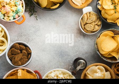Snacks. Ungesunde Lebensmittel. Alle klassischen Kartoffelsnacks mit Erdnüssen, Popcorn- und Zwiebelringen und gesalzenen Brezeln in Schüsseln auf grauem Beton-Hintergrund Stockfoto