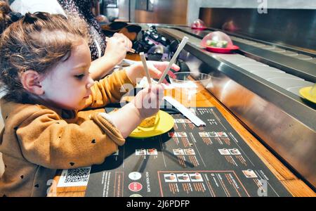 Nettes kleines Mädchen, das in einem Restaurant mit Selbstbedienungsförderband Essstäbchen zum Sushi verwendet Stockfoto