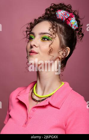 Porträt einer jungen lächelnden Frau in rosa Kleid auf rosa Hintergrund gekleidet. Weibchen mit ungewöhnlichen grünen Augen Schatten Make-up, lockiges Haar und Ohrringe. Stockfoto