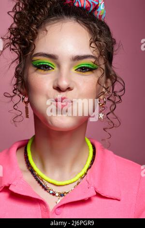 Mädchen pufft ihre Wangen heraus. Porträt einer jungen Frau in rosa Kleid auf rosa Hintergrund gekleidet. Weibchen mit ungewöhnlichen grünen Augen Schatten Make-up, lockiges Haar Stockfoto