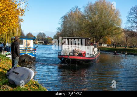 Großes, schmal gefahrenen Boot und Kanus auf dem River Cam mit einem Mann, der am Flussufer neben dem Windsurfer in Cambridge, England, steht Stockfoto