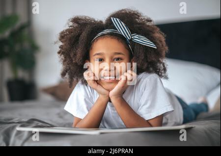 Ein süßes Mädchen mit Haarband, das mit einem Buch auf dem Bett liegt Stockfoto