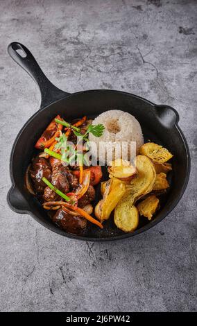 Peruanisches Essen - Lomo saltado - Rinderfilet mit lila Zwiebeln, gelber Paprika, Tomaten in einer schwarzen Pfanne mit pommes frites und Reis serviert. Draufsicht Stockfoto