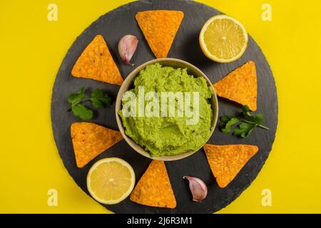 Zusammensetzung von Guacamole mit den Zutaten für seine Kreation und die Nachos für seinen Verzehr auf einem gelben Hintergrund. Stockfoto