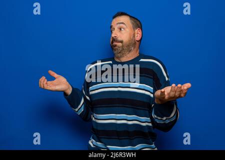 Lateinischer bärtiger Mann, gekleidet in einen hässlich gestreiften Pullover mit den Handflächen nach oben und mit den Händen in Resignation-Geste, isoliert auf blauem Hintergrund Stockfoto