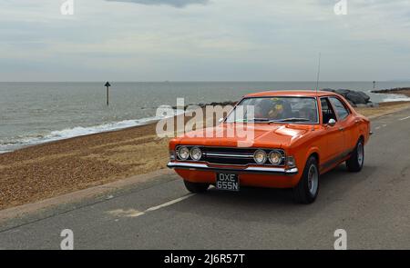 Classic Orange Ford Cortina MK3 Auto, das entlang der Strandpromenade und dem Meer im Hintergrund gefahren wird. Stockfoto