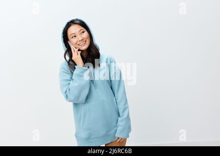 Schöne glücklich hübsche koreanische junge Frau in blauen Hoodie halten Telefon Gespräche mit Freund posiert isoliert auf über weißem Studio Hintergrund. Das beste Angebot Stockfoto