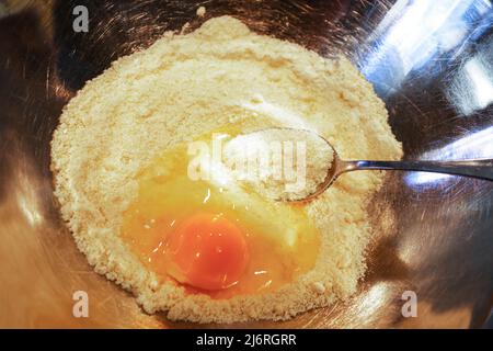 Glutenfreie kochende kochende Speisen - ein Ei, das in einen Haufen Mandelmehl zerbrochen ist, mit einem Löffel, der bereit ist, es in einer Edelstahlschüssel aufzurühren - Nahaufnahme Stockfoto