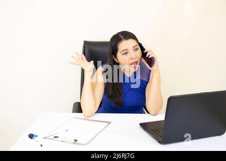 Der nette, erfolgreiche CEO-Assistent mit blauer Bluse überraschte den Anruf auf dem Mobiltelefon, dem Schreibtisch mit Ducuments und dem Laptop auf weißem Hintergrund Stockfoto