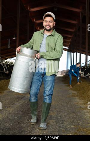 Landwirt, der im Kuhstall Milch trägt, in der Nähe des Stalls mit Kühen Stockfoto