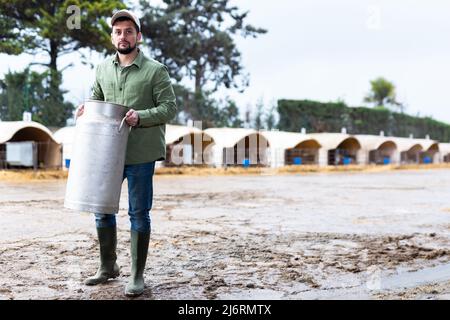 Der männliche Landwirt trägt eine große Milchkrüge in seinen Händen auf dem Kuhbetrieb Stockfoto