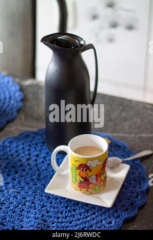 Frühstück auf einem Granittisch mit einem weißen quadratischen Teller und einer bunt gemusterten Tasse Latte, Brotscheiben mit Erdnussbutter Stockfoto
