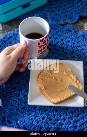 Frühstück auf einem Granittisch mit einem weißen quadratischen Teller und einer bunt gemusterten Tasse Latte, Brotscheiben mit Erdnussbutter Stockfoto