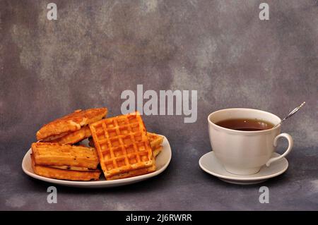 Ein Teller mit mehreren frischen belgischen Waffeln mit Creme und einer Tasse schwarzem Tee auf grauem Hintergrund. Nahaufnahme. Stockfoto