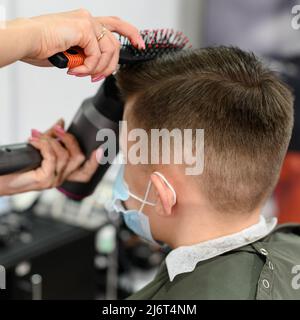 Teenager bekommt einen Haarschnitt während einer Pandemie in einem Friseurladen, einen Haarschnitt und trocknet Haare nach einem Haarschnitt, styling Haare nach einem Haarschnitt mit einem Haartrockner. Stockfoto