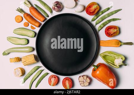 Kreative Gestaltung von Weißen, Tomaten, Paprika, Karotten, Knoblauch, Mais, Zucchini, Zwiebel und schwarze Platte gemacht. Bio Lebensmittel Hintergrund. Flach, Ansicht von oben, Stockfoto