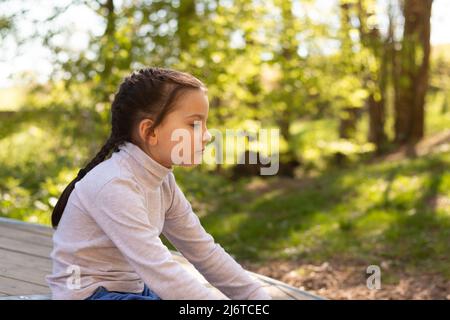 Eine kleine, schön lächelnde Schülerin auf einem natürlichen Hintergrund in einem Rollkragenpullover und einer blauen Jeans sitzt auf dem Boden, umarmt ihre Beine und schaut nach Aw Stockfoto