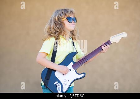 Kleiner Junge spielt Gitarre im Freien. Kindermusiker spielt Gitarre wie ein Rockstar im Freien. Kid Boy Rockmusiker mit Gitarre. Musik für Kinder. Stockfoto