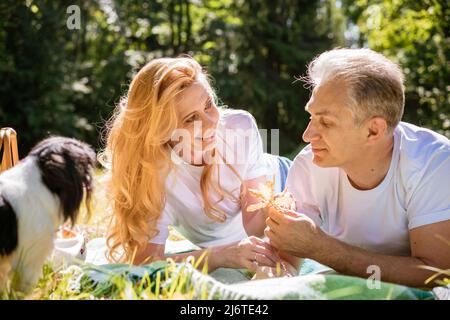 Reifes Paar liegt mit ihrem Hund im Park. Ein älteres Paar ruht in der Natur mit Hund. Nahaufnahme eines älteren Mannes und einer älteren Frau in weißen Hemden und Jeans. Stilvolle und moderne Großeltern. Stockfoto