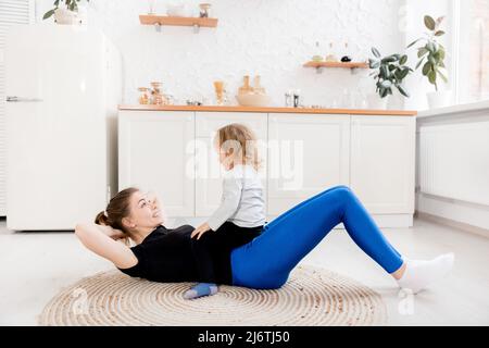 Am Morgen geht die lächelnde Mutter mit ihrer kleinen Tochter in die Küche zum Sport. Gesundes Lifestyle-Konzept. Schulung während Stockfoto