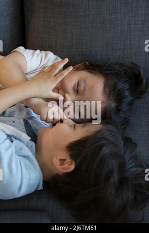 Ein kleines Mädchen und ein kleiner Junge liegen kopfüber auf dem Sofa und kneifen sich gegenseitig ins Gesicht - Stockfoto Stockfoto