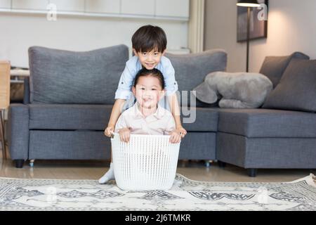 Am Sofa im Wohnzimmer war ein kleines Mädchen bereit, im Wäschekorb zu sitzen und der kleine Junge versuchte, sie nach oben zu bewegen Stockfoto