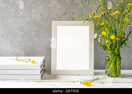 Modellerstellung. Weißer Rahmen auf weißem Holztisch mit grauem Betonhintergrund, Büchern, Notizbuch und wilden Blumen Stockfoto
