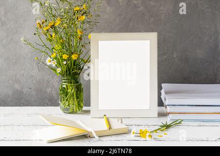 Modellerstellung. Weißer Rahmen auf Holztisch mit grauem Betonhintergrund, Büchern, Notizbuch, Bleistift und Wildblumen Stockfoto