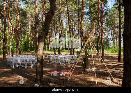 Wunderschöne böhmische Tipi-Bogendekoration auf der Hochzeitsfeier im Freien im Pinienwald mit Kegeln. Stühle, floristische Blumenkompositionen aus Rosen Stockfoto