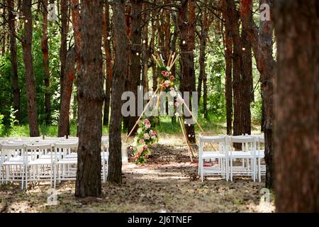 Wunderschöne böhmische Tipi-Bogendekoration auf der Hochzeitsfeier im Freien im Pinienwald mit Kegeln. Stühle, floristische Blumenkompositionen aus Rosen Stockfoto