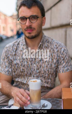 Junger brauner bärtiger ernster Mann mit einem Latte Macchiato-Milchcofee auf einem Tisch, der das Getränk in einem Hemd mit kurzen Ärmeln Bergamo Shop Italy packt Stockfoto