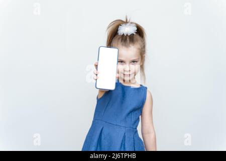 Ich kann es empfehlen. Kleines Mädchen in blauem Kleid mit leerem Handy-Bildschirm, grauer Studiowand Stockfoto