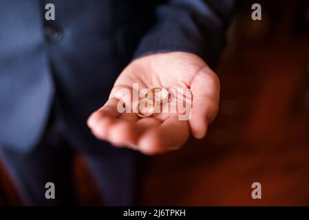 Auf der Handfläche eines Mannes befinden sich zwei Eheringe. Der Bräutigam hält die Ringe. Hochzeitstradition. Goldene Ringe liegen auf der Handfläche eines Mannes Stockfoto