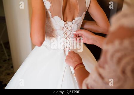 Die Brautjungfer hilft der Braut, das weiße Hochzeitskleid zu kleistchen. Die Hände und Knöpfe der Mutter auf dem Brautkleid in Nahaufnahme. Die Braut setzt sich auf sie Stockfoto