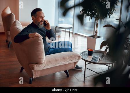 Glücklicher bärtiger Mann, der neben dem Fenster saß und sich am Telefon unterhielt Stockfoto