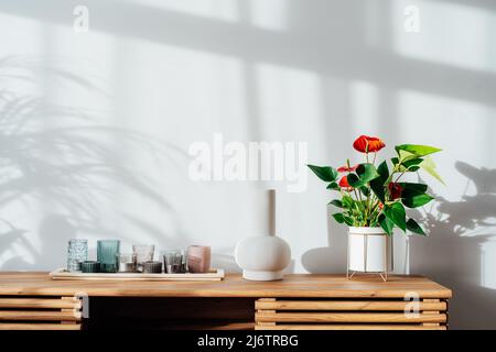 Moderne Einrichtung im minimalistischen skandinavischen Stil. Kerzen, Keramikvase und House Pflanzen rotes Anthurium in einem Topf auf einer Holzkonsole unter Sonnenlicht und Sha Stockfoto