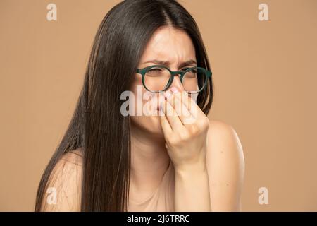 Porträt einer frustrierten Frau, die sich wegen des schlechten Geruchs auf beigem Hintergrund mit Ekel in die Nase kneift. Stockfoto