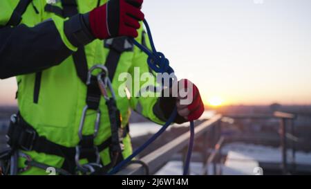 Industriekletterer im grünen Outfit bindet Knoten mit Schlaufe an, der Karabiner am Sicherheitsseil befestigt und am Geländer befestigt wird. Der Mann bereitet sich darauf vor, aus der Nähe die Wand des Wolkenkratzers hinunterzuklettern Stockfoto