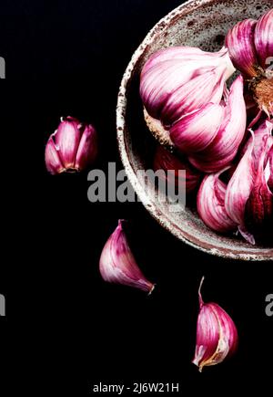 Frischer reifer roter Knoblauch in Keramikschale und einige Nelken auf schwarzem Hintergrund Stockfoto