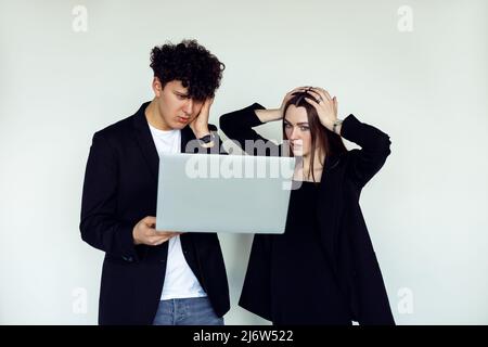 Porträt eines jungen überemotionalen Paares in schwarzer Kleidung. Besorgter Mann, der einen Laptop in der Nähe einer schockierten Frau hält, die den Kopf greift. Stockfoto