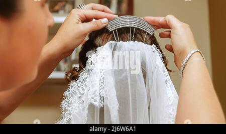 Das silberne Haarzubehör aus Strass wird von den Händen einer Frau in das Haar einer Braut gelegt, während sie sich an ihrem Hochzeitstag kleidet. Stockfoto