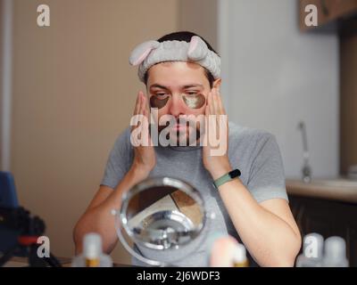 Augenflecken. Junger Mann in der Küche mit Stirnband auf dem Kopf und Spiegel Anwendung unter Augen Anti-Falten-Patches, Männer machen morgens Gesichtsbehandlungen mit Hydrogel Beauty-Produkt, Hautpflege. Stockfoto