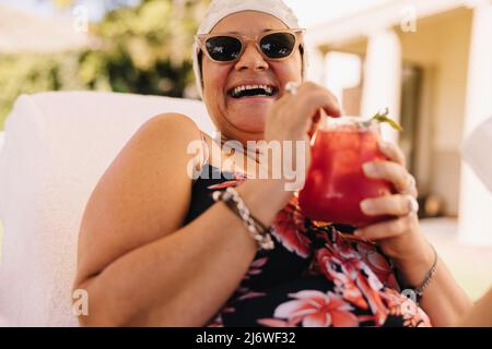 Glückliche ältere Frau, die während des Urlaubs im Sommer einen Cocktail genießt. Sorglose ältere Frau, die fröhlich lacht, während sie sich auf einer Liege entspannt. Reif Stockfoto