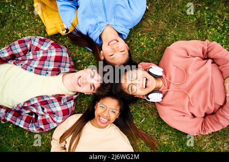 Selbstbewusste multirassische Gruppe von Studenten, die Bücher halten und die Kamera lächeln, die im Kreis auf dem Rasen liegt. Fröhliche junge Menschen, die Spaß haben Stockfoto