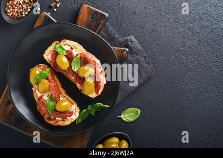 Bruschetta mit sonnengetrockneten Tomaten. Toast oder Sandwich mit Ricotta, sonnengetrockneten Tomaten und Provence-Kräutern mit Basilikum auf schwarzem Teller auf schwarzem Teller gezackt Stockfoto