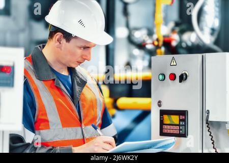 Der junge Ingenieur, der Helm und Schutzweste verwendet, zeichnet die Messwerte der Geräte oder des Gasverbrauchs im Kesselraum auf. Beginn der Heizperiode und Gasversorgung. Stockfoto