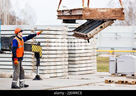 Slinger in Helm und Weste steuert das Entladen von Metallkonstruktionen auf der Baustelle. Weißer Handwerker entlädt Ladung. Stockfoto