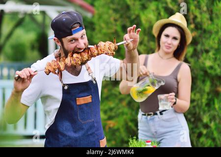 Der junge bärtige Mann in einer Mütze und einer Jeanschürze lächelt und hält einen Spieß mit köstlichem Grillfleisch, das vor dem Hintergrund auf dem Grill zubereitet wird Stockfoto