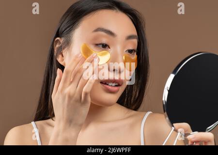 Junge asiatische Dame hält Spiegel und Anwendung unter Augenflecken Maske, Herstellung von Schönheitsbehandlungen, brauner Hintergrund Stockfoto